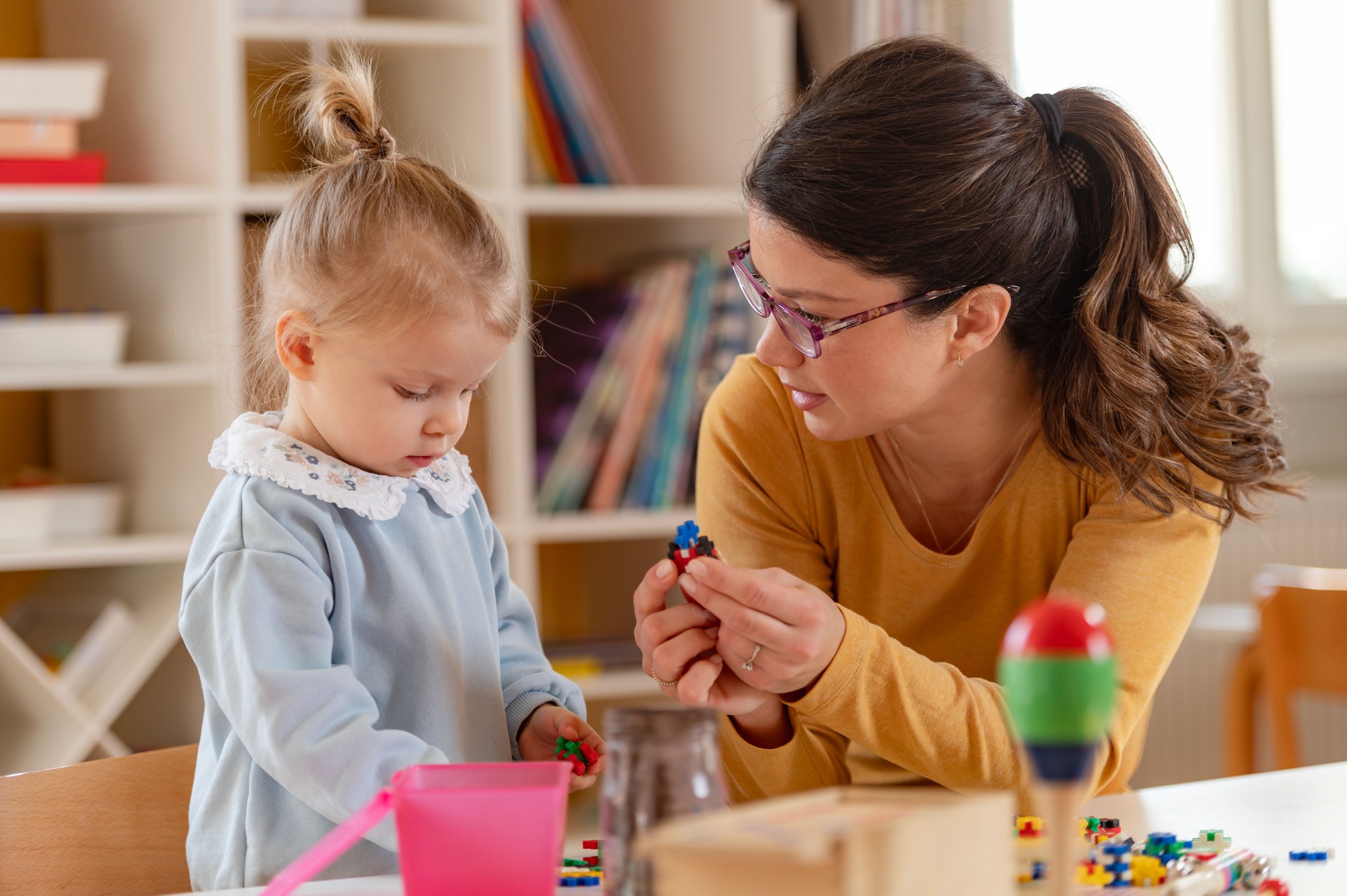 A smiling woman and a learning toddler girl explore sensory and educational toys together in a bright classroom.